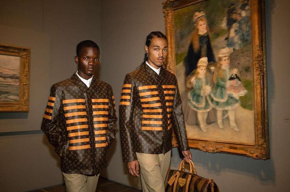 Two Black men in designer jackets pose inside an art gallery next to a framed painting, holding a patterned handbag with leather trim.