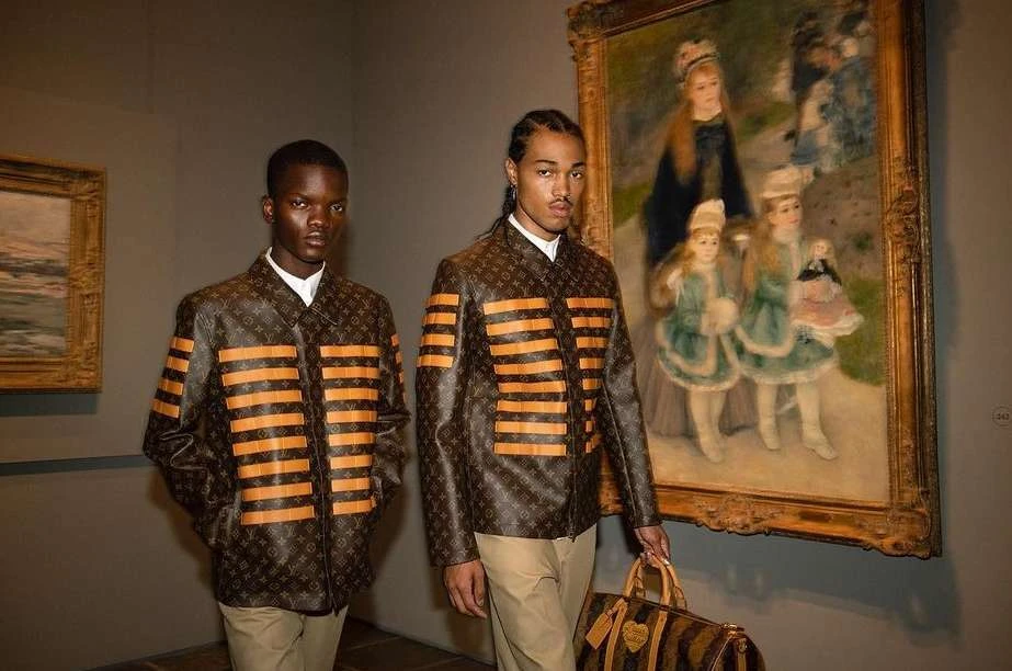 Two Black men in designer jackets pose inside an art gallery next to a framed painting, holding a patterned handbag with leather trim.