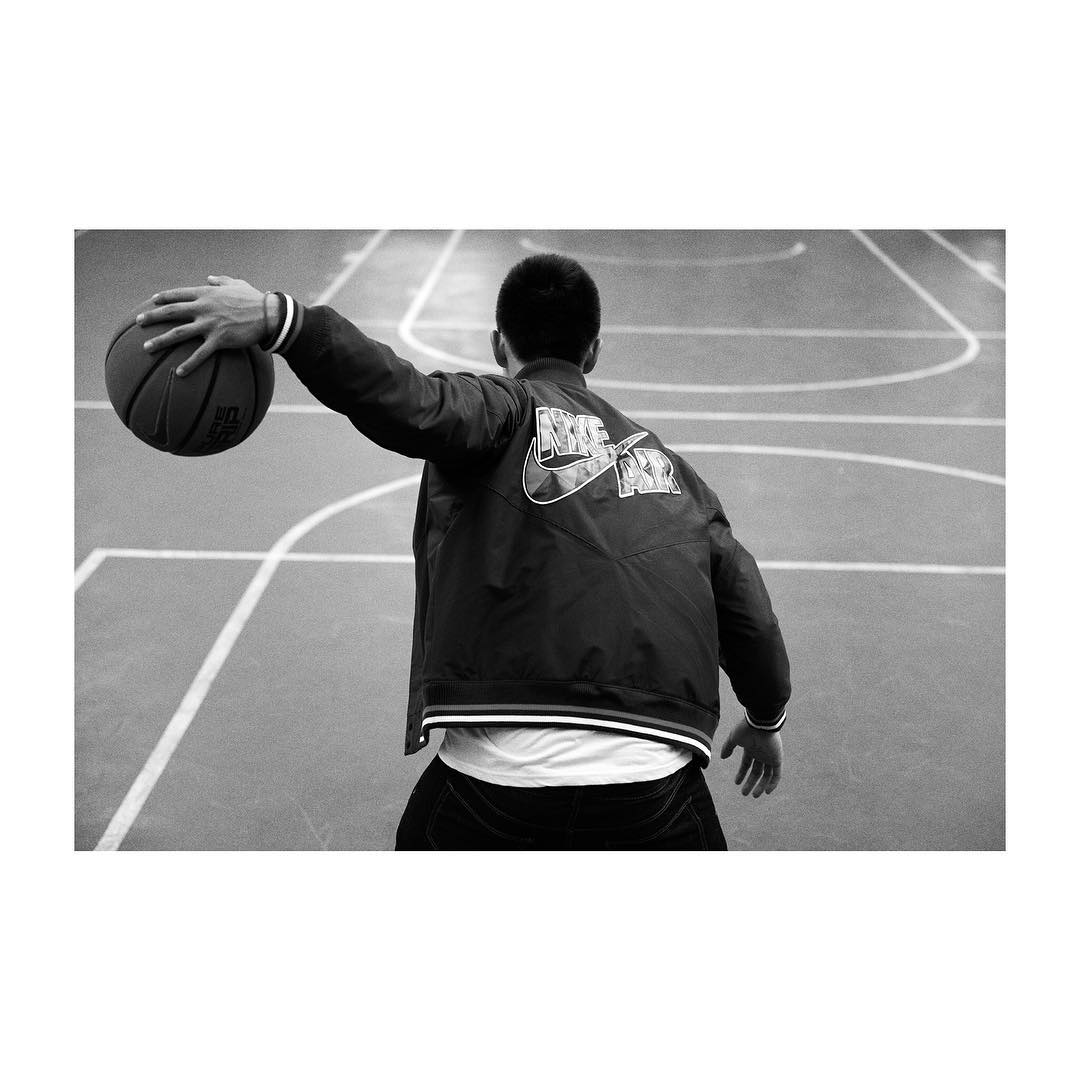 Back view of a man in a Nike jacket holding a basketball on an indoor court, monochrome photography, indoor sports fashion.