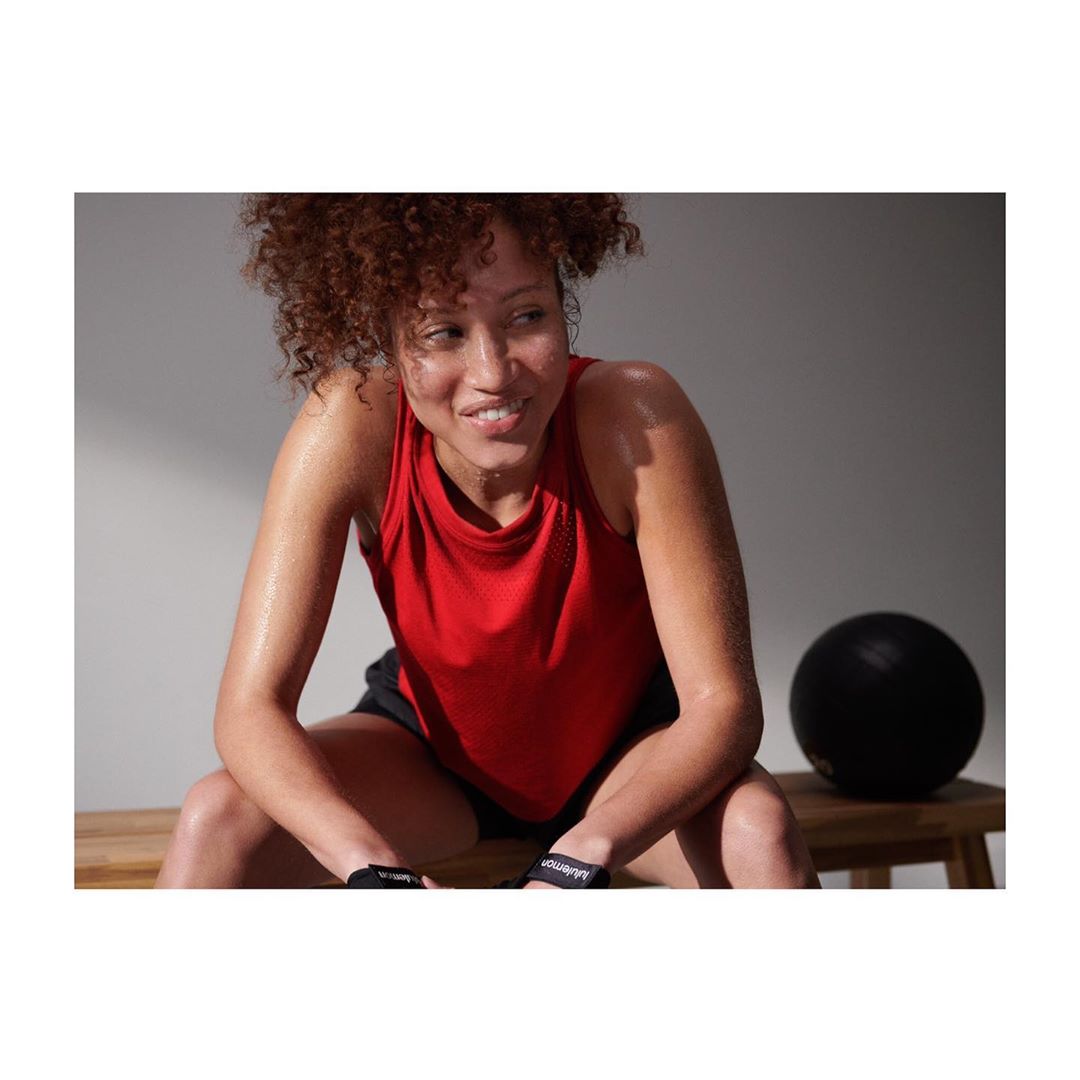 Athletic woman with curly hair in workout clothes sitting and smiling in a gym setting with a medicine ball in the background.