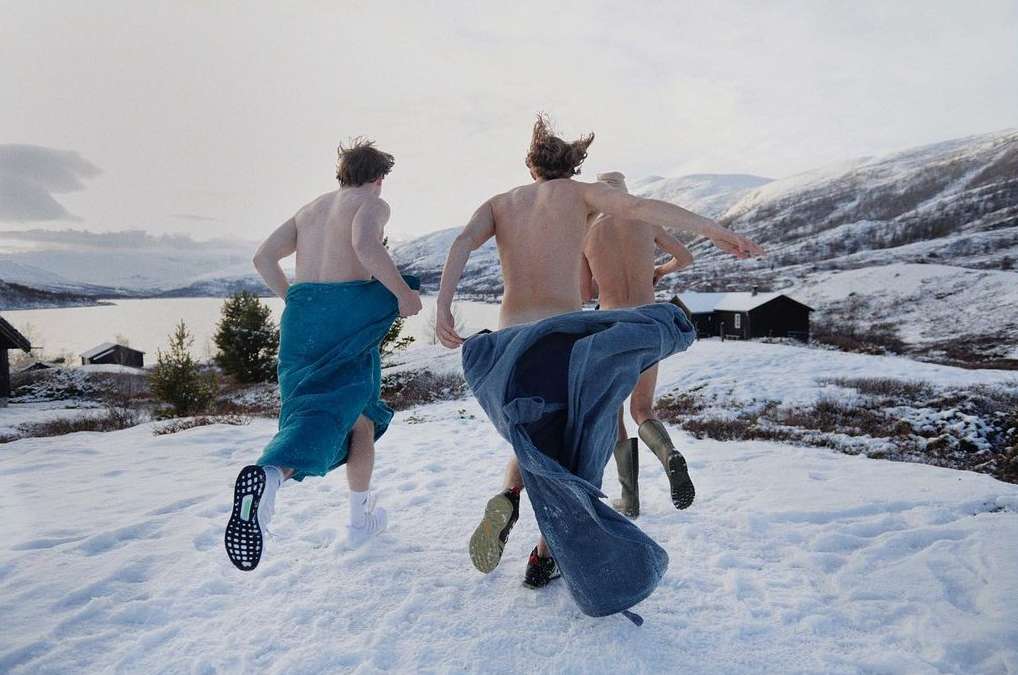Three shirtless men run in snow covered landscape near a lake, wearing boots and towels, mountain backdrop under overcast skies.