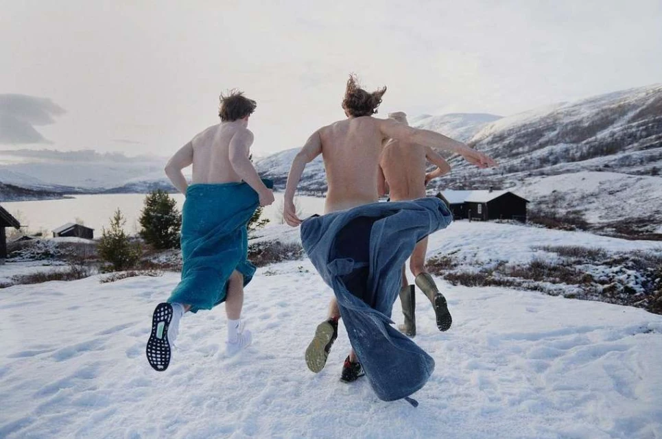 Three shirtless men run in snow covered landscape near a lake, wearing boots and towels, mountain backdrop under overcast skies.