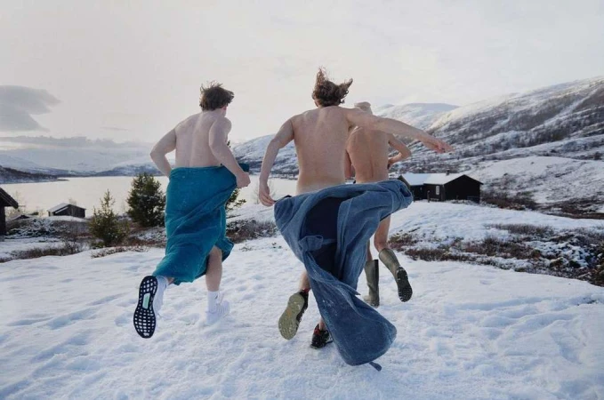Three shirtless men run in snow covered landscape near a lake, wearing boots and towels, mountain backdrop under overcast skies.
