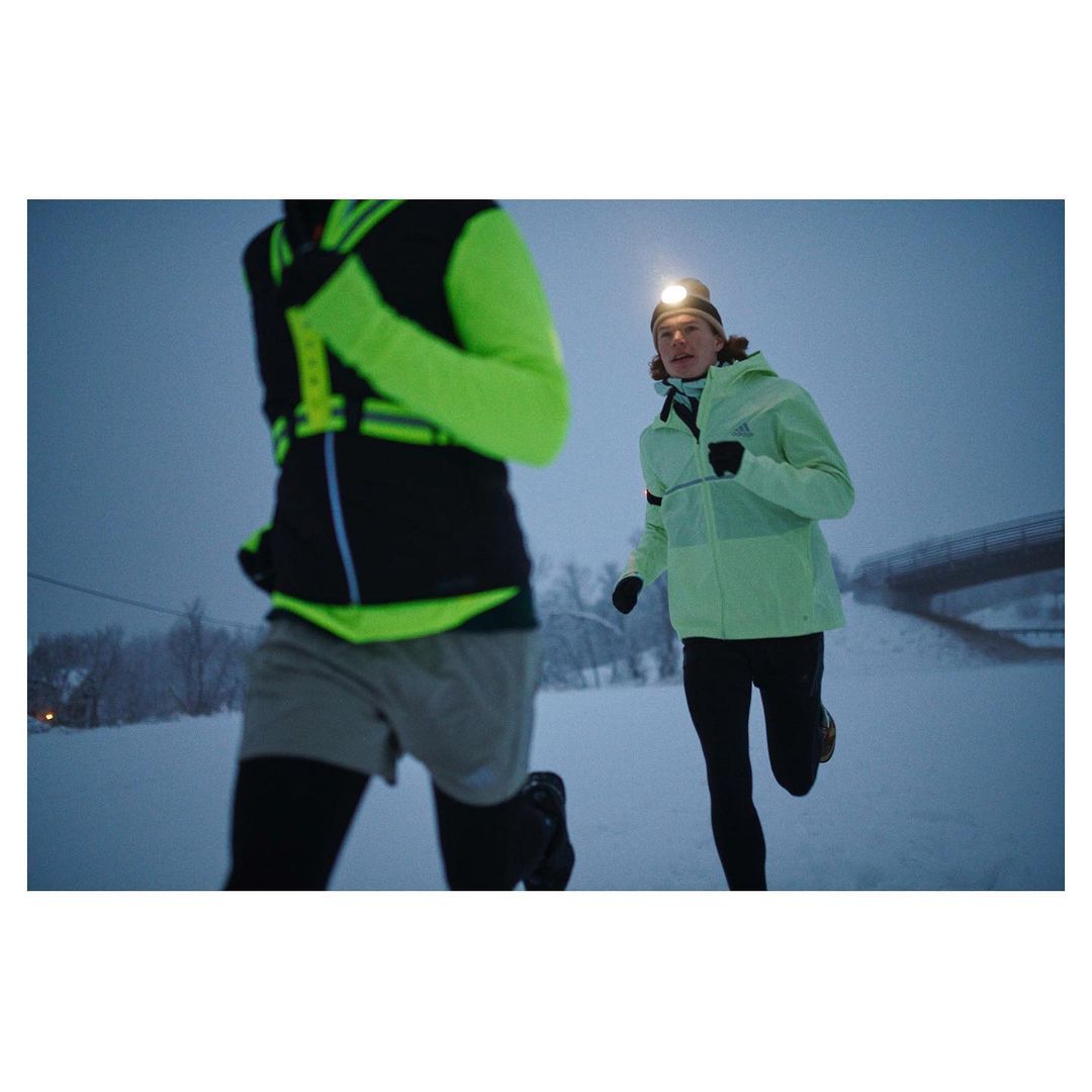 Two people running in a snowy outdoor environment. The person in the foreground has a headlamp. Both are wearing athletic clothing.
