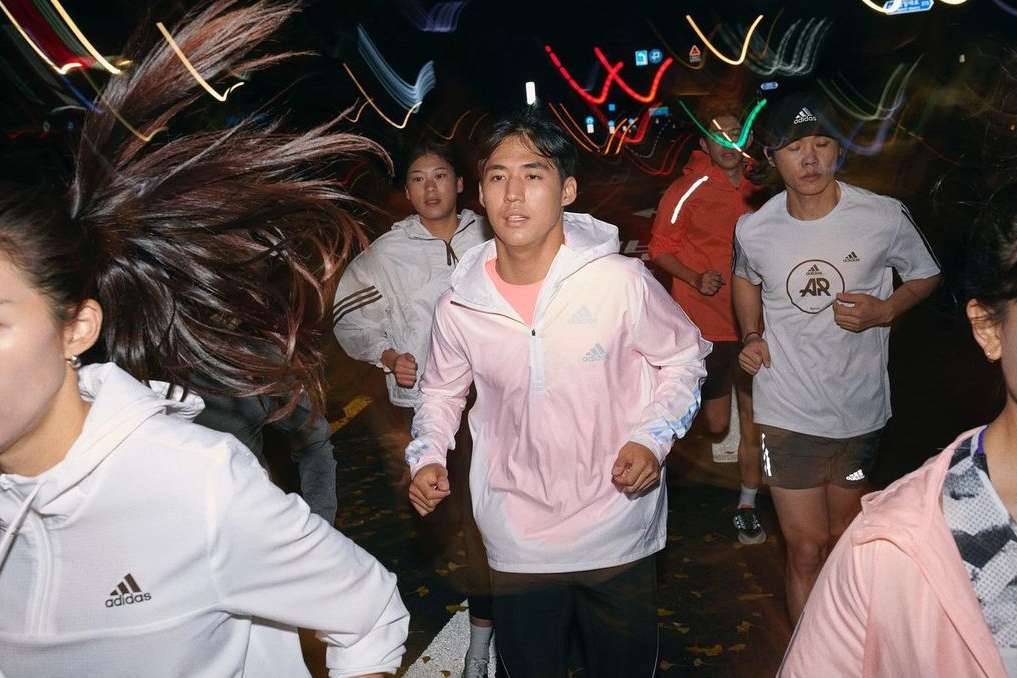 Group of people running at night on city street. Focus on Asian adult male wearing pink Adidas windbreaker and black pants.