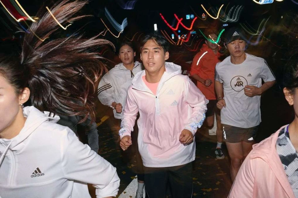 Group of people running at night on city street. Focus on Asian adult male wearing pink Adidas windbreaker and black pants.