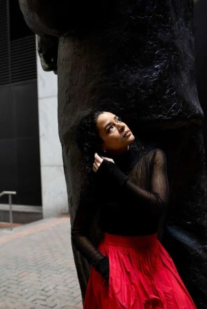 Woman in a sheer black top and red skirt poses next to a sculpture outdoors, looking upward in a medium shot with a brick background.