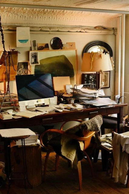 Cluttered artist's workspace with a computer, papers, desk lamp, and art supplies, creating a busy and creative atmosphere.
