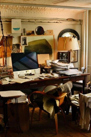 Cluttered artist's workspace with a computer, papers, desk lamp, and art supplies, creating a busy and creative atmosphere.