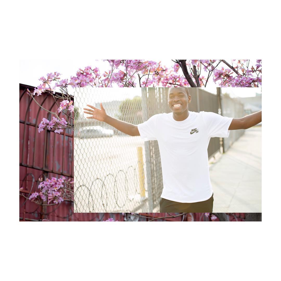 Man in white t-shirt smiles with open arms near pink flowering tree against wire fence and urban street during daylight.