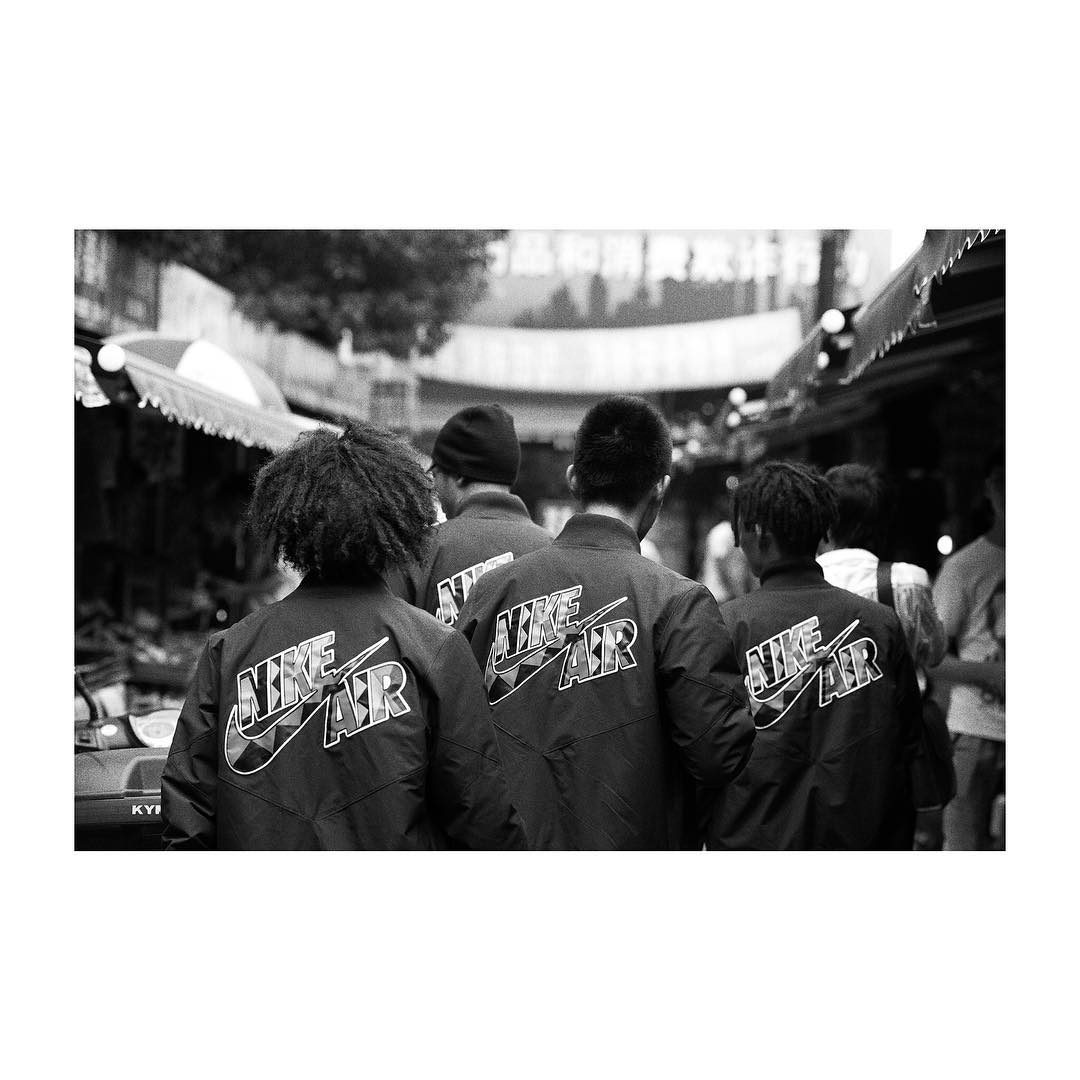 Four people wearing matching Nike Air jackets walk away in a monochrome urban setting with market stalls and building details visible.
