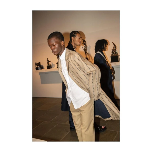 Three male fashion models pose in a museum setting, showcasing a striped suit jacket, white shirt, and tan pants in soft lighting.