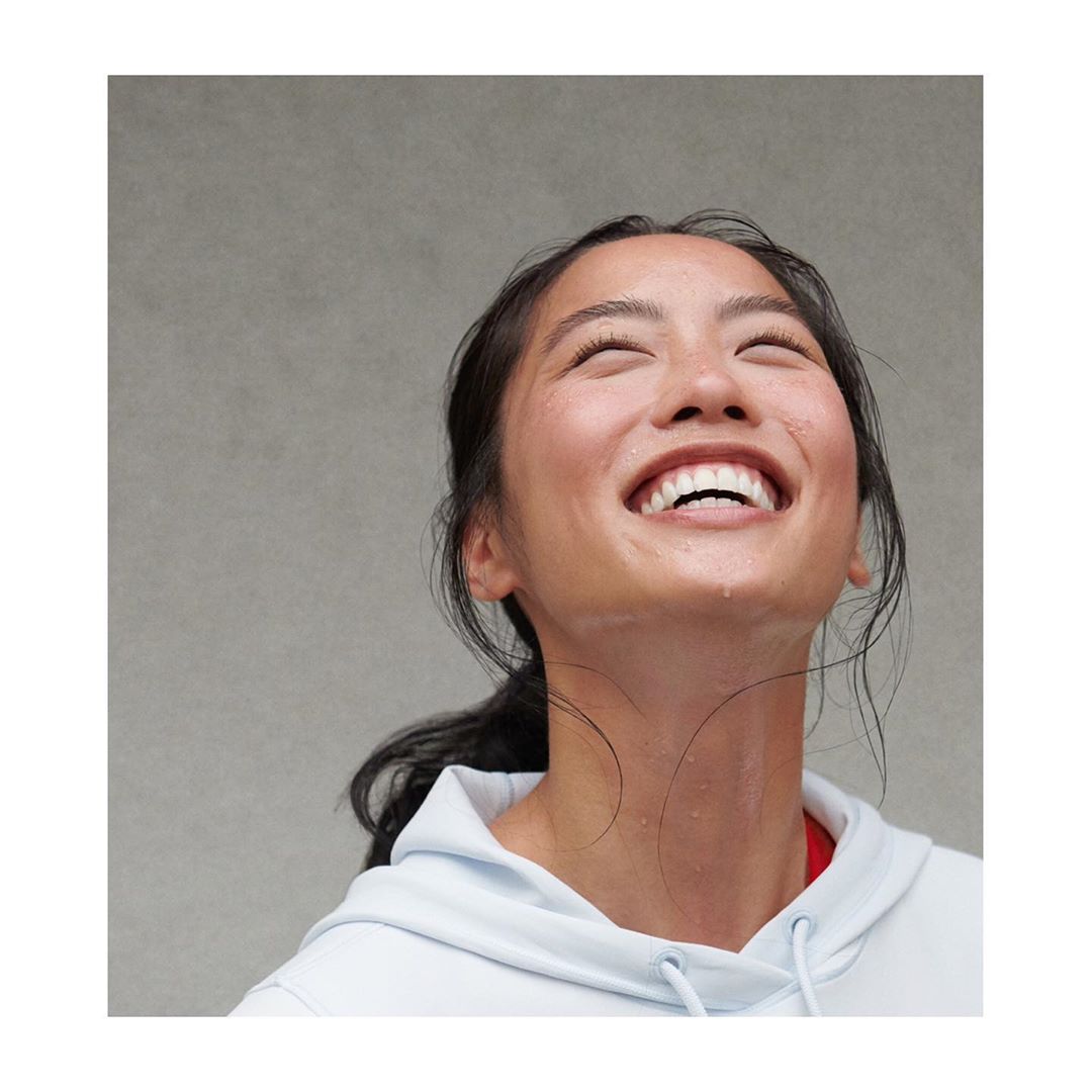 Asian woman looking up with a bright smile. She has sweat on her face and neck. She's wearing a light blue sweatshirt with a red shirt underneath.