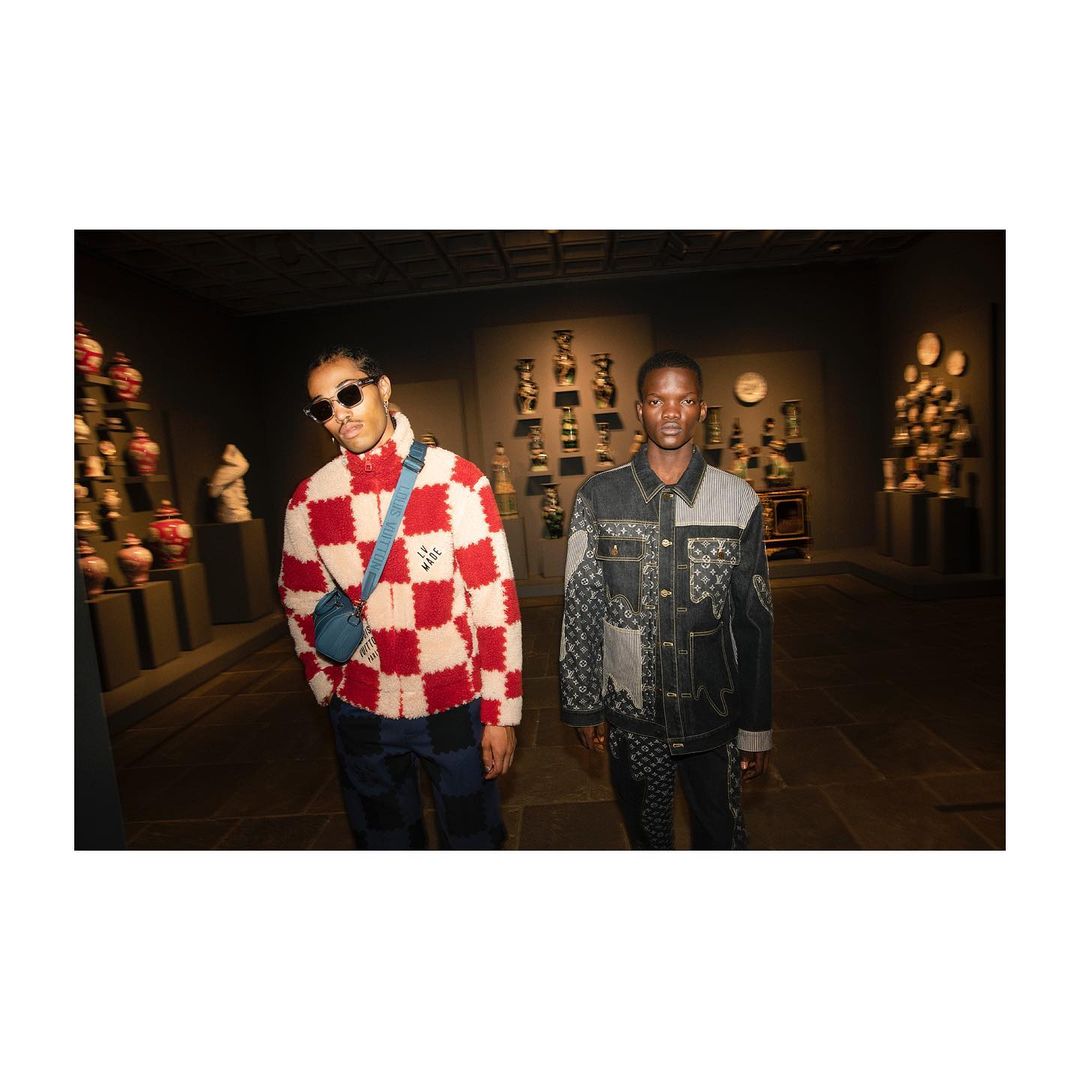Two Black men pose in a museum-like setting, one in a red-and-white jacket with a blue bag, and the other in a patchwork denim jacket.