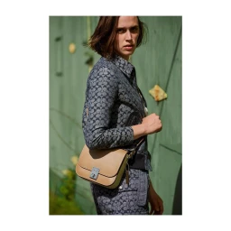 Woman in patterned denim jacket and pants holding a tan leather shoulder bag outdoors against a green wall