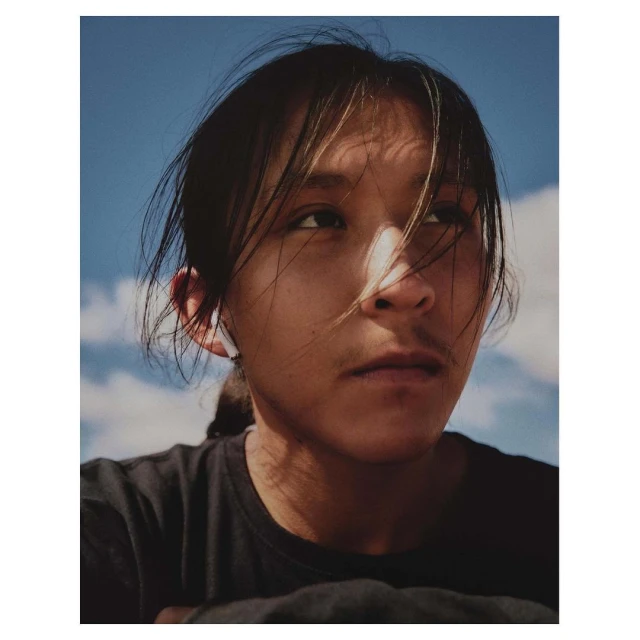 Close-up portrait of a young adult with dark hair against a blue sky with clouds, creating a serene and contemplative mood.