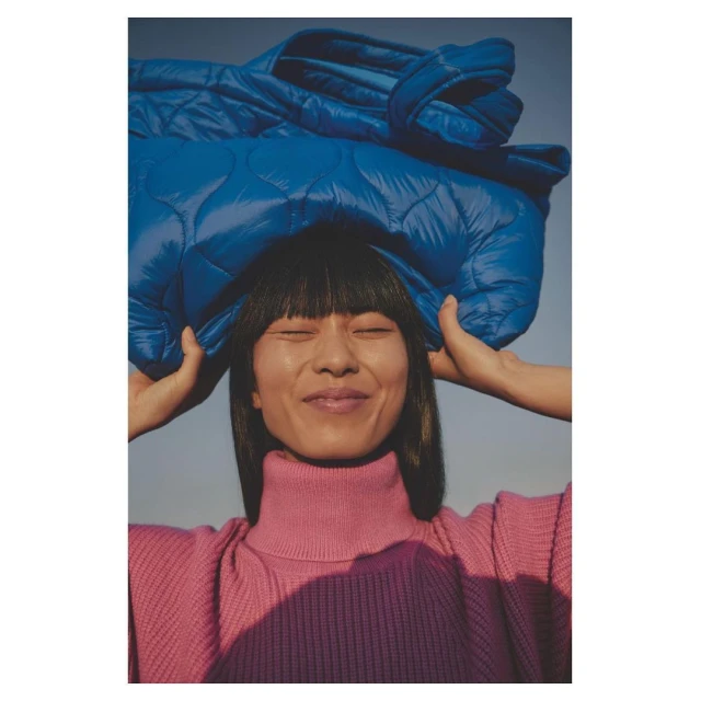 Woman in pink turtleneck smiles with eyes closed while holding a folded, quilted, blue jacket over her head in an outdoor setting.