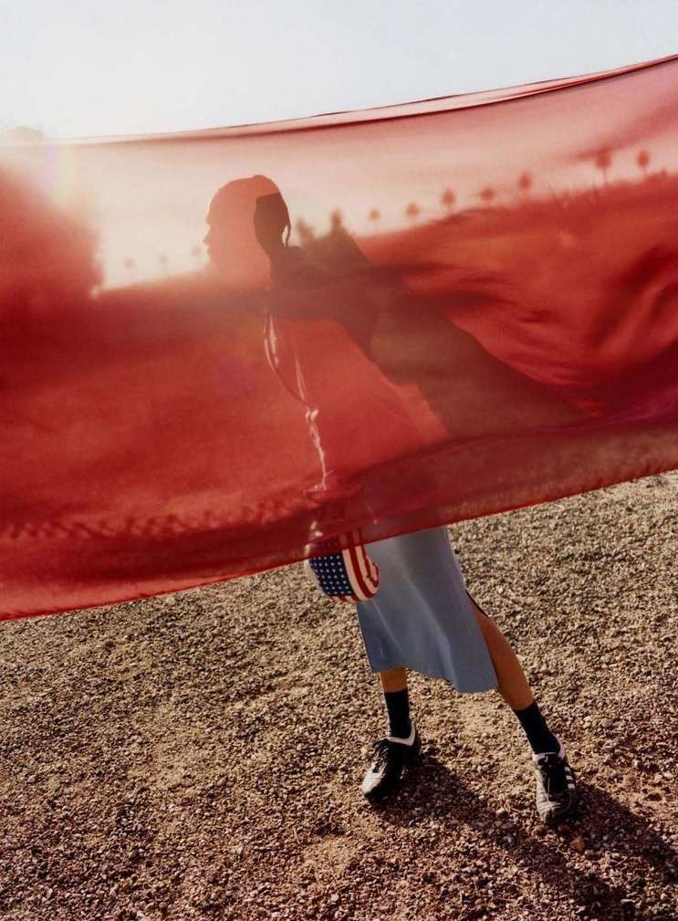 Woman in motion with American flag on gravel surface. She is wearing a blue dress with black shoes and socks and a red transparent fabric.