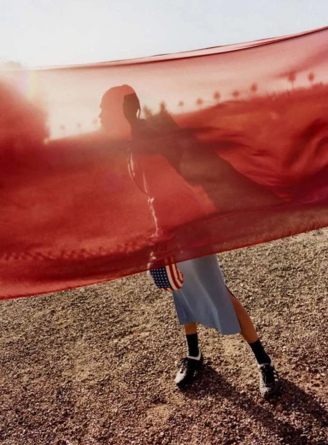 Woman in motion with American flag on gravel surface. She is wearing a blue dress with black shoes and socks and a red transparent fabric.