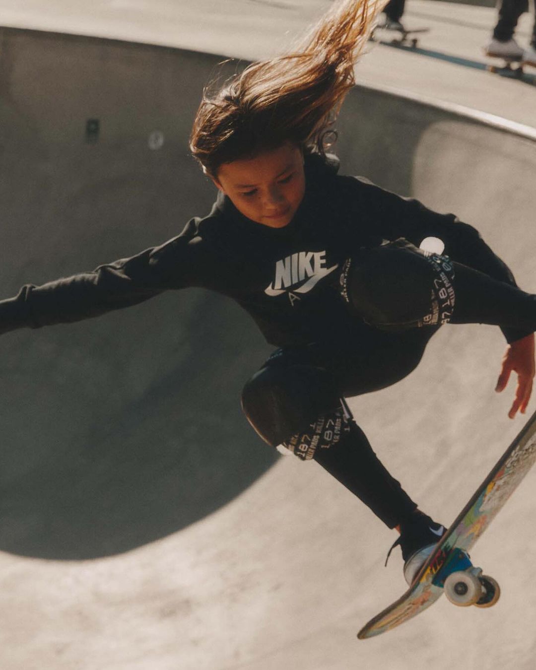 Young skateboarder soars in a concrete skatepark, wearing Nike sportswear, catching air on a skateboard, and showing movement and style.