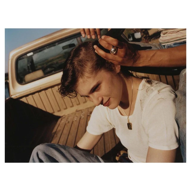 Woman getting a haircut outdoors in a pickup truck bed. A second person holds hair above her head in this sunlit, medium shot.