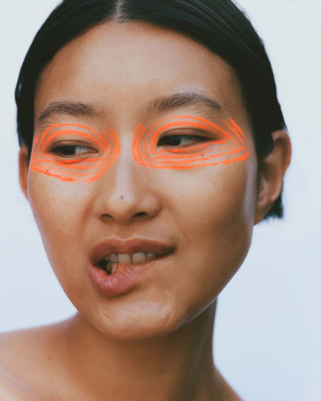 Headshot of an East Asian adult female with orange paint around her eyes, light complexion, dark hair, and a white background.
