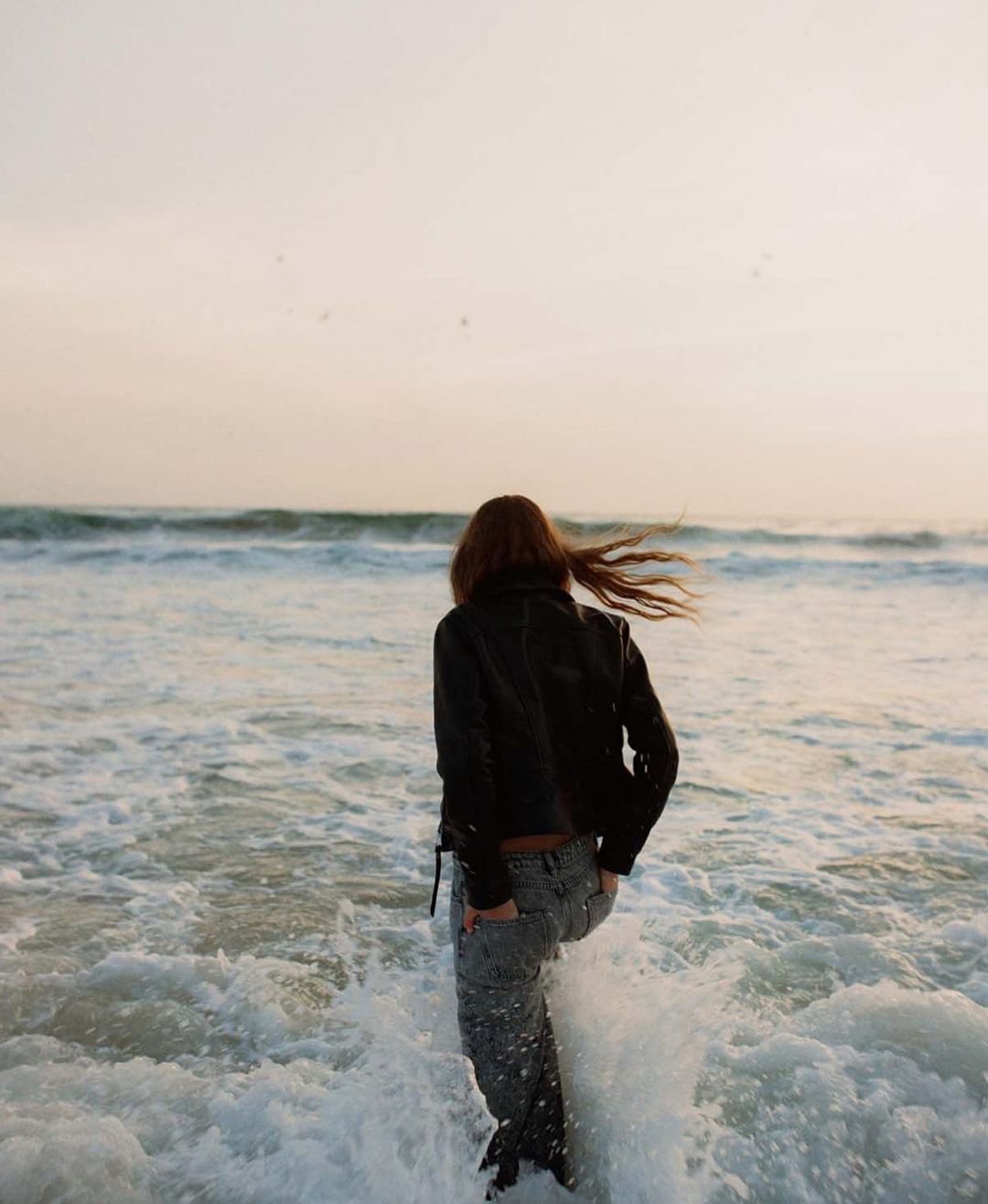 Woman in black leather jacket and jeans walks into ocean waves at sunset. Back view of female figure in water at the beach.