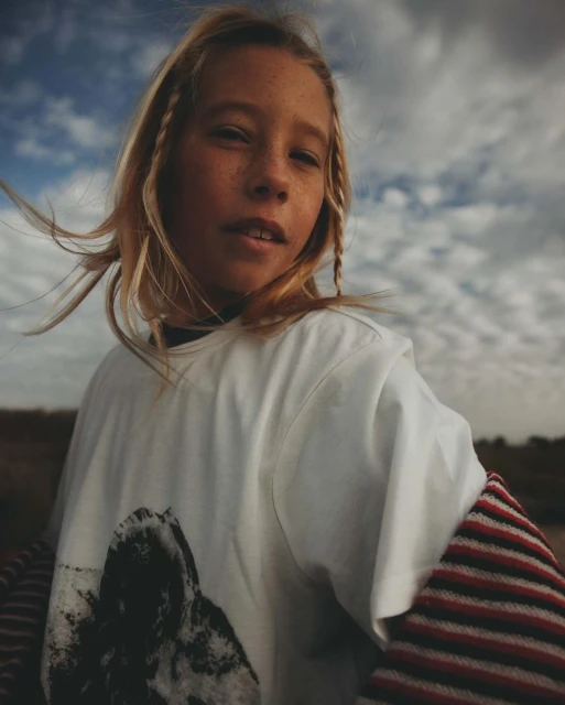 Portrait of a child with blonde braids, wearing a white tee with a graphic print, standing outdoors with a cloudy sky in the background.