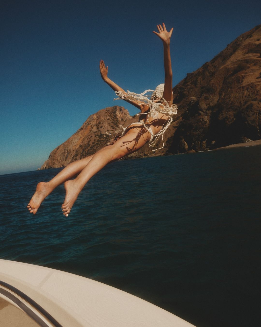 Woman in swimsuit jumps from a white boat into the ocean with mountains in the background, captured under a clear blue sky.