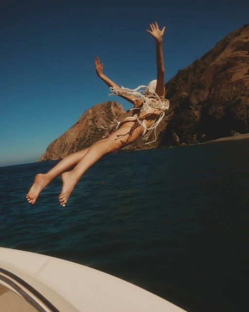 Woman in swimsuit jumps from a white boat into the ocean with mountains in the background, captured under a clear blue sky.