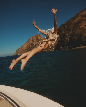 Woman diving mid-air into deep blue ocean water with rocky coastal cliffs and clear blue sky in background