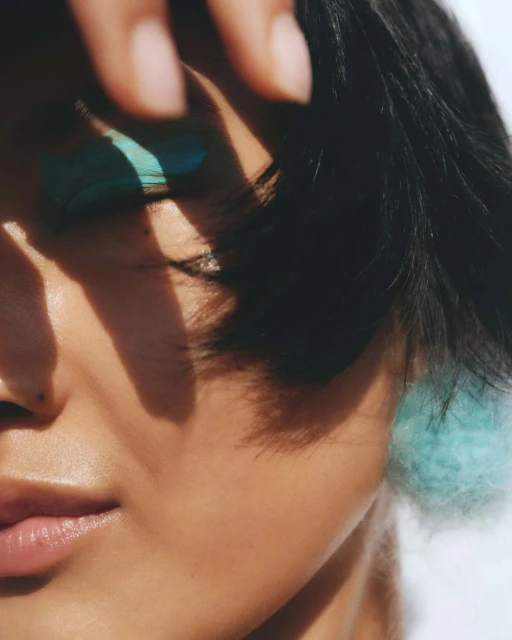 Close-up portrait of an adult woman with short black hair wearing blue eye shadow and a fluffy blue earring, with shadows on her face.
