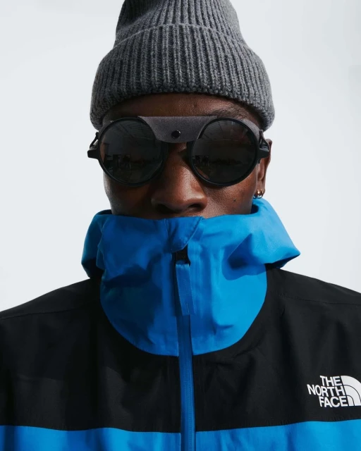 Close-up of a stylish Black man wearing a beanie, unique sunglasses, and a blue and black North Face jacket against a white background.