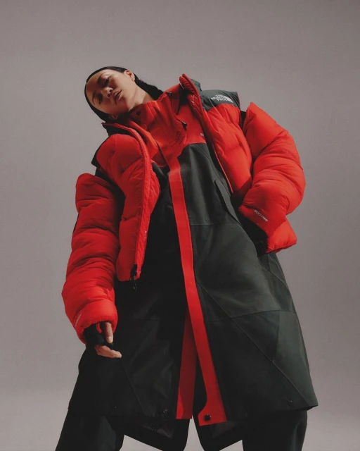 Portrait of a person wearing a bright red down jacket and dark overall-like garment against a soft grey studio backdrop with diffused lighting.