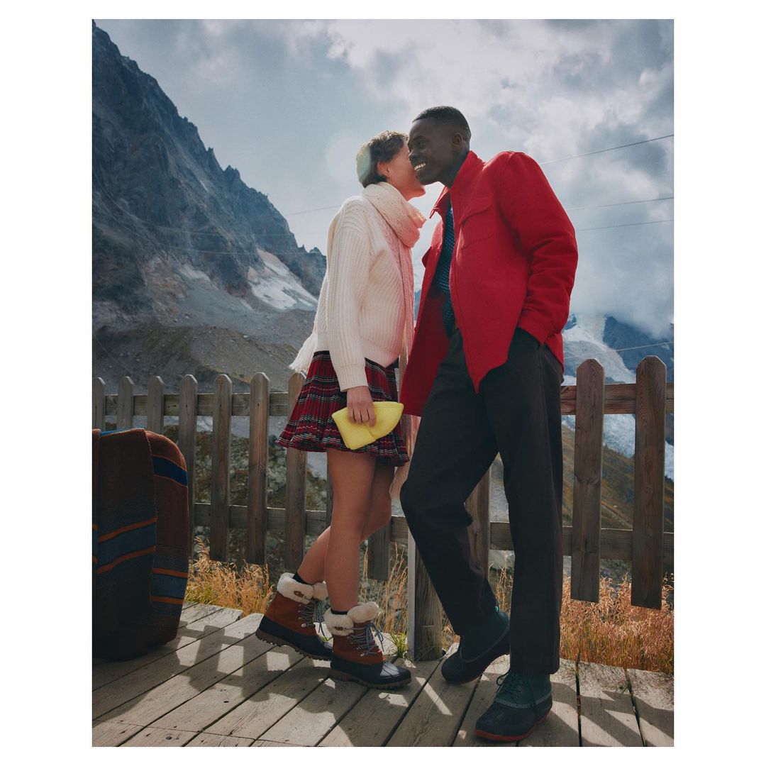 A smiling African-American man and Caucasian woman in winter clothes stand on a wooden deck against a scenic mountain backdrop.