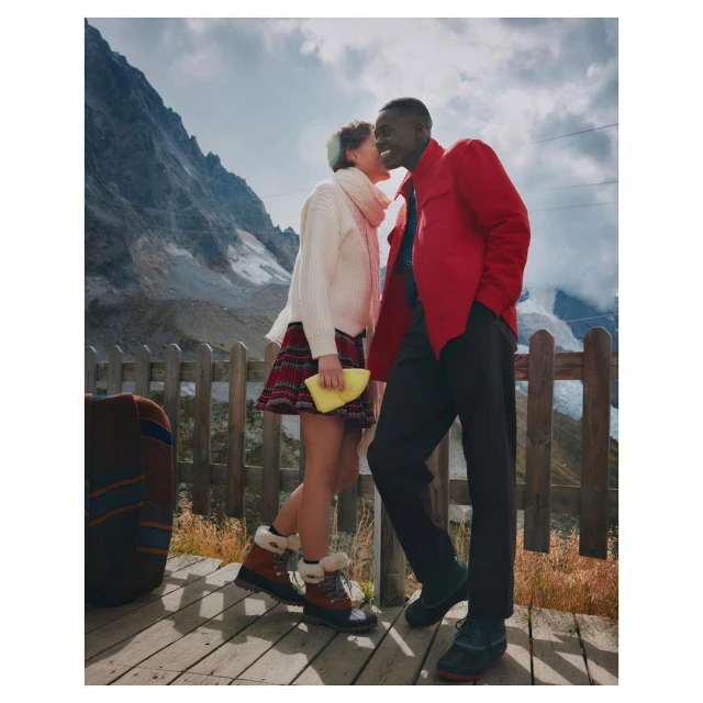 A smiling African-American man and Caucasian woman in winter clothes stand on a wooden deck against a scenic mountain backdrop.