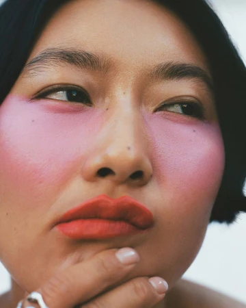 Close-up portrait of a woman with vibrant pink blush and orange lipstick, resting chin on hand.
