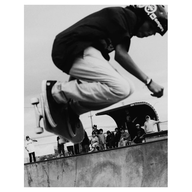 Black and white shot of a skateboarder in mid-air performing a trick, with a group of onlookers and a skatepark visible in the background.