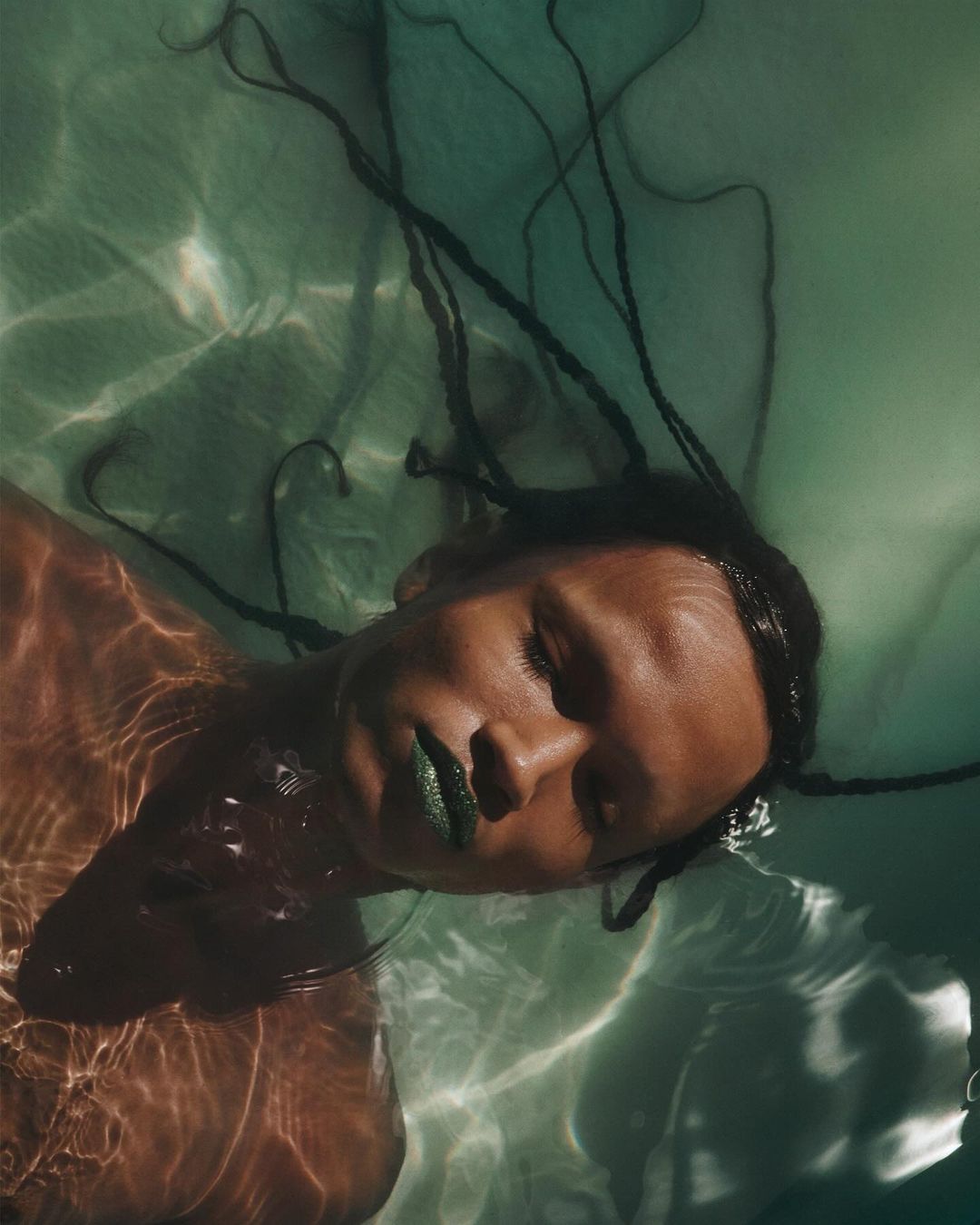 Woman with brown skin and long braids floats in rippled, light green water, wearing green lipstick in a portrait orientation.