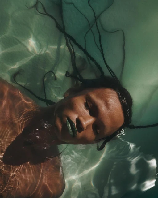 Woman with brown skin and long braids floats in rippled, light green water, wearing green lipstick in a portrait orientation.