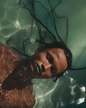 Woman with brown skin and long braids floats in rippled, light green water, wearing green lipstick in a portrait orientation.