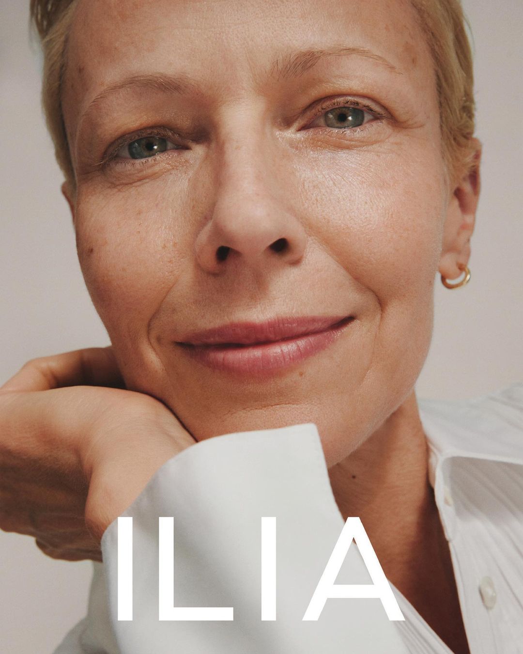 Close-up of a smiling fair-skinned woman with short blonde hair wearing a white button-down shirt against a light background.