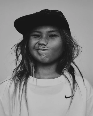 Portrait of a child with long hair, wearing a black cap and white Nike t-shirt, making a funny face in a studio shot.