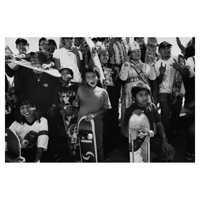 Black and white photo of a group of young people, some holding skateboards, with one in ceremonial dress, captured at eye-level.