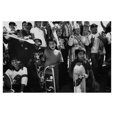 Black and white photo of a group of young people, some holding skateboards, with one in ceremonial dress, captured at eye-level.