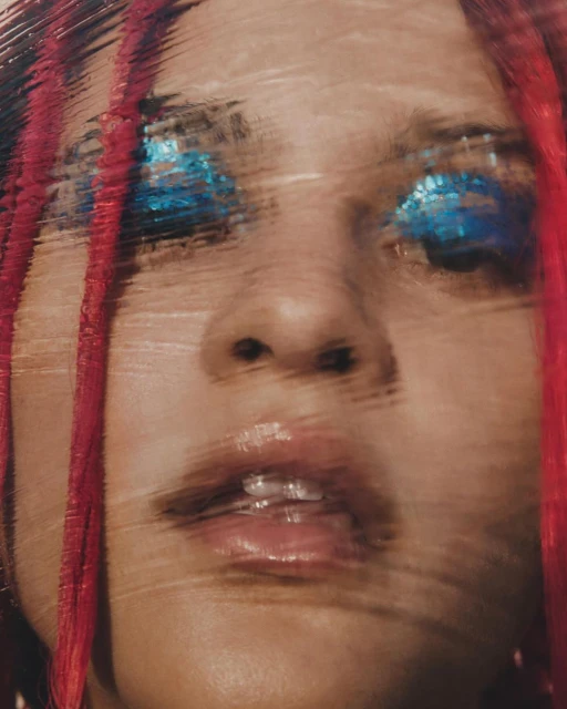 Close-up portrait of an adult female with dramatic blue eye makeup, red braids, and a blurry, textured overlay effect for a striking visual.