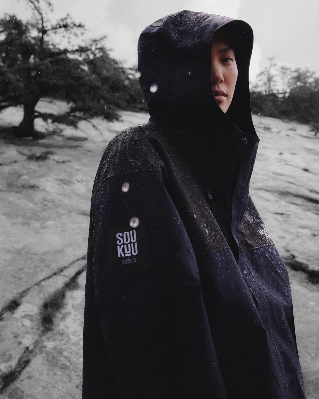 Portrait of a person wearing a dark hooded waterproof jacket with a Soukuu logo patch outdoors in a rocky, overcast landscape.