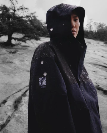 Portrait of a person wearing a dark hooded waterproof jacket with a Soukuu logo patch outdoors in a rocky, overcast landscape.