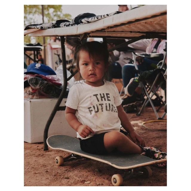Young girl with pigtails sits cross-legged on a skateboard wearing a white 'The Future' T-shirt in an outdoor dirt environment.