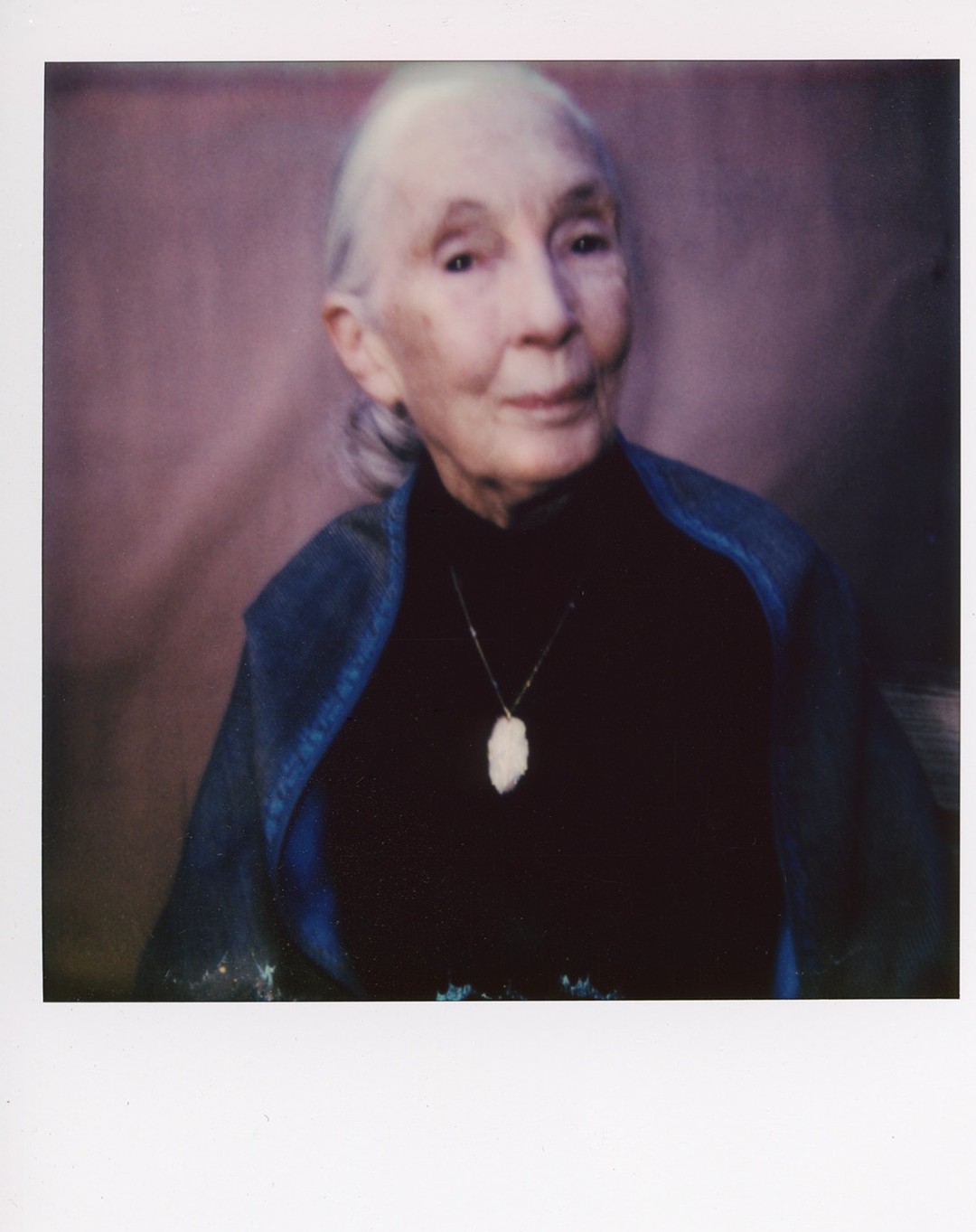 Portrait of an elderly adult woman in a blue jacket and black turtleneck, wearing a pendant necklace, against a mottled pink background.